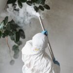 A person in protective gear uses a sprayer for indoor disinfection.